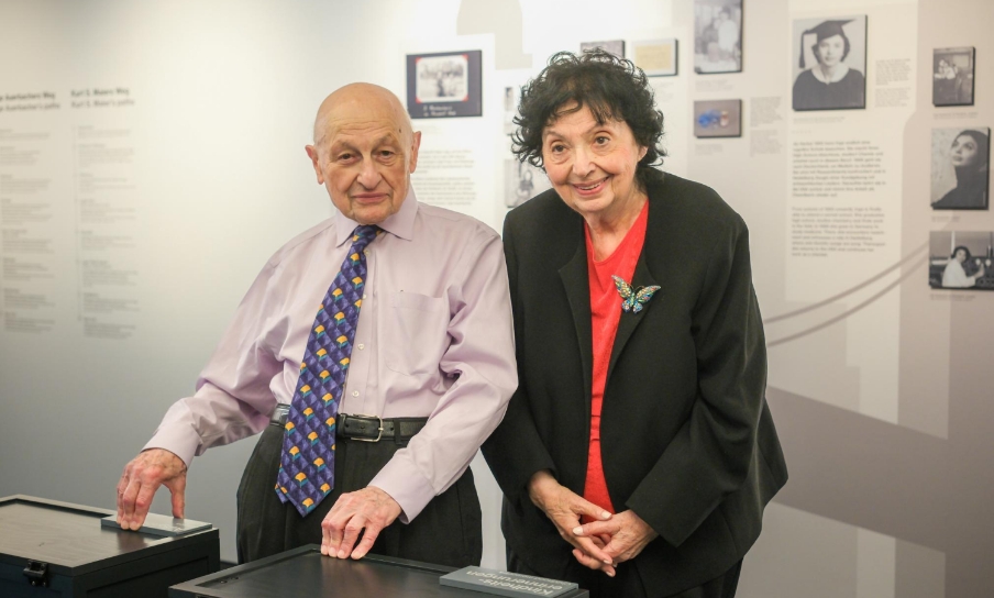 Zwei Zeitzeug*innen in einer Ausstellung: Mann mit Hemd und bunter Krawatte, Frau in Businesskleidung mit rotem Oberteil vor einer Infowand.