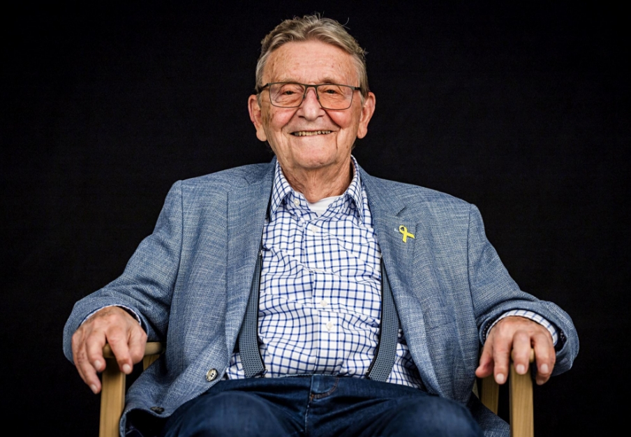 Älterer Zeitzeuge lächelt im Sessel vor dunklem Hintergrund, mit blauem Sakko, kariertem Hemd und Hosenträgern.